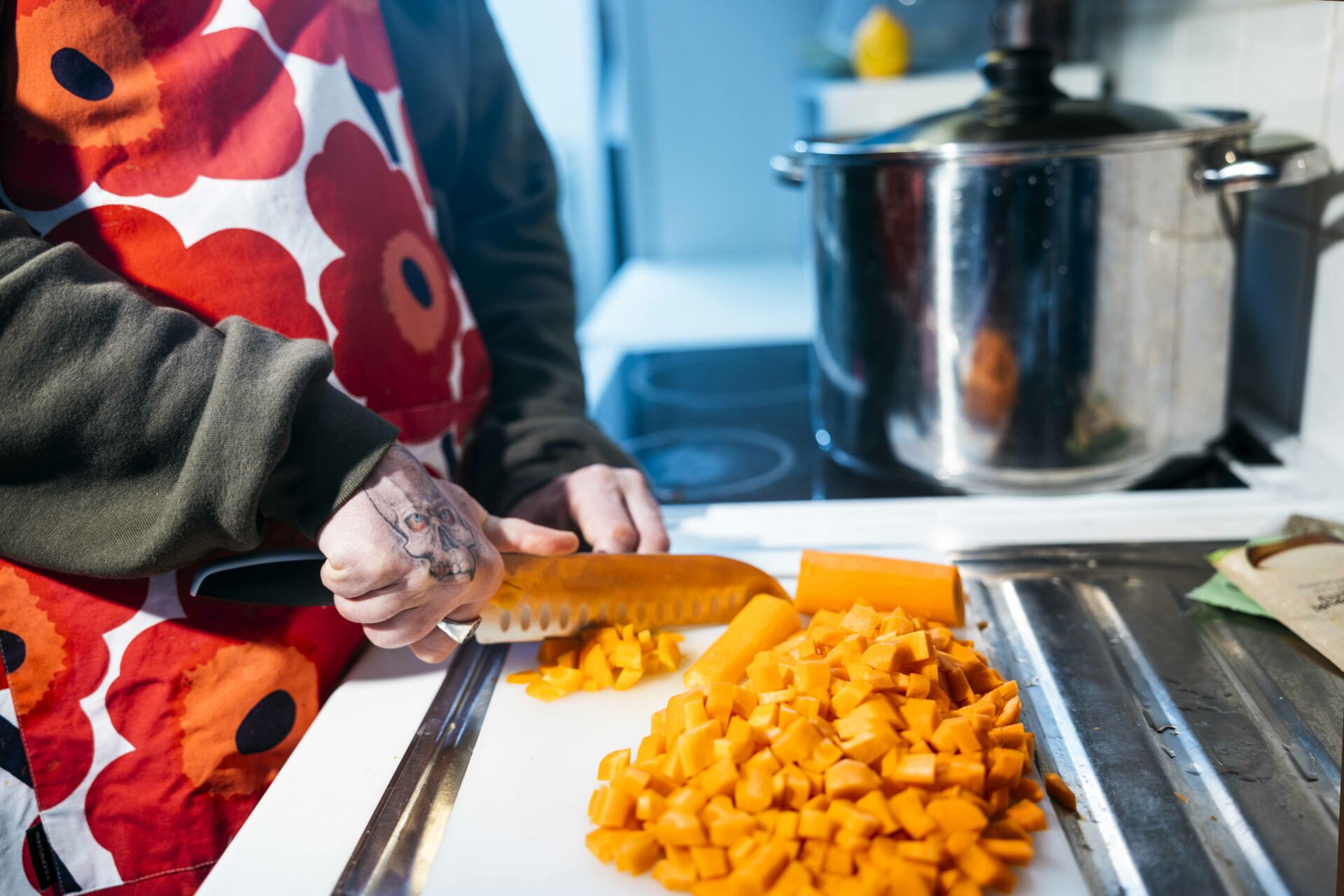 Mies leikkaa porkkanoita keittiössä. Hänellä on unikko-essu ja tatuointi kädessään.